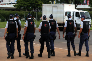 Eine Gruppe von Beamten in Uniform, einschließlich Mützen, Hemden, Schutzwesten, Uhren, Hosen und Schuhen, die auf einer Straße mit einem geparkten Lastwagen auf der rechten Seite neben einem Zaun, Gebäuden, Bäumen, Pflanzen, Pfählen und Bannern im Hintergrund und einer Brücke im oberen rechten Eck gehen.