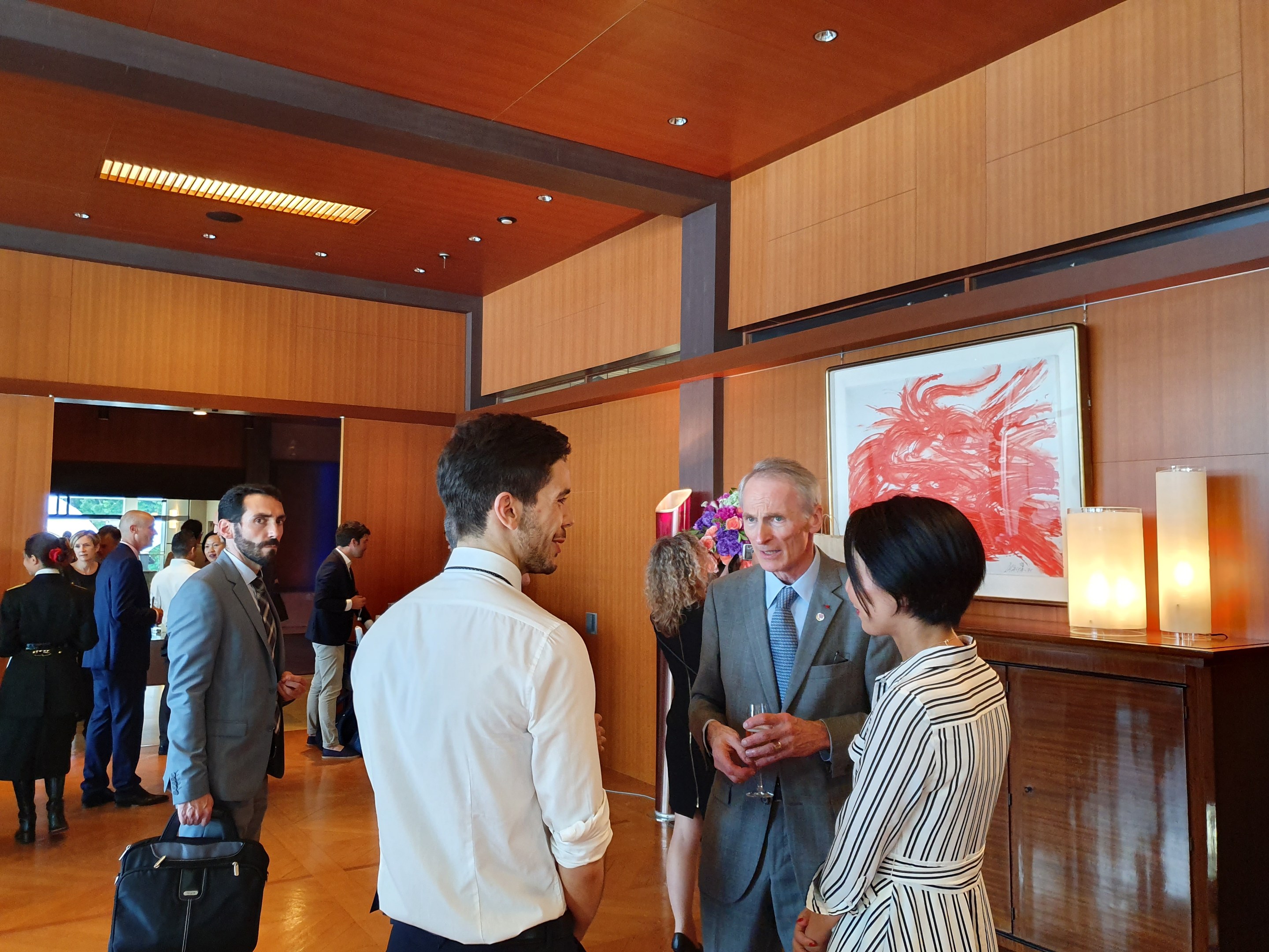 Menschen, die in einem Raum mit Holzverkleidung und einem Foto Rahmen sowie Beleuchtung stehen und sich unterhalten, wobei einige Taschen tragen, auf einer von der Nationalen Kunstgalerie Singapurs veranstalteten Kunstmesse.