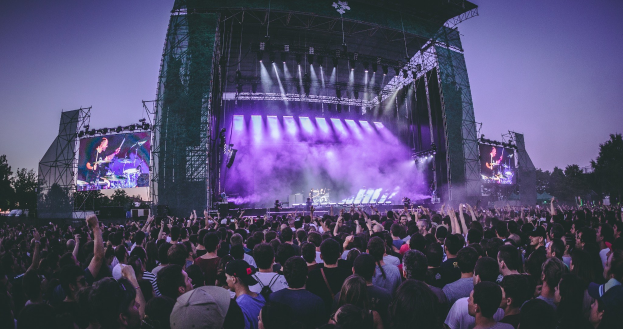 Eine große Menschenmenge bei einem Musikfestival schaut auf einer beleuchteten Bühne mit Instrumenten und Bildschirmen, die vor einem Gebäude, Bäumen und einem blauen Himmel aufgebaut sind.