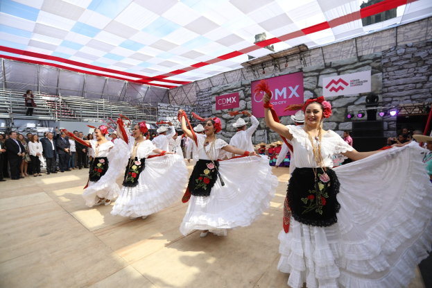 Eine Gruppe von Frauen in weißen Kleidern tanzt auf einer Bühne, umgeben von einer Zuschauermenge, einem Videografen auf der rechten Seite und Bühnenbeleuchtung und Texttafeln im Hintergrund.