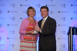 Zwei Personen, die eine Trophäe halten, wobei eine Person einen schwarzen Blazer und ein weißes Hemd trägt und ein Banner im Hintergrund zu sehen ist.