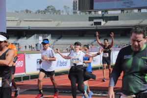 Gruppe von Menschen, die auf einer Stadionbahn laufen, mit Bannern, einer Anzeigetafel, einem Gebäude, Bäumen und einem bewölkten Himmel; einige Läufer tragen Turnschuhe und Hüte.