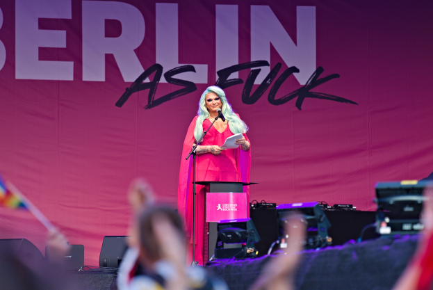 Eine Frau in einem pinken Kleid steht an einem Podium mit einem Mikrofon und Papier, spricht zu einer Menge mit einem 'Berlin as Fuck'-Plakat im Hintergrund.