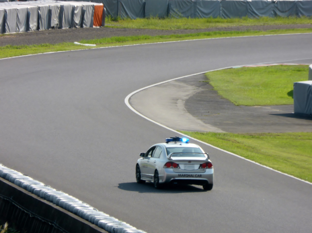 Polizeiauto fährt auf einer Rennstrecke mit Gras auf beiden Seiten, mit Menschen, Zelten und anderen Gegenständen im Hintergrund.