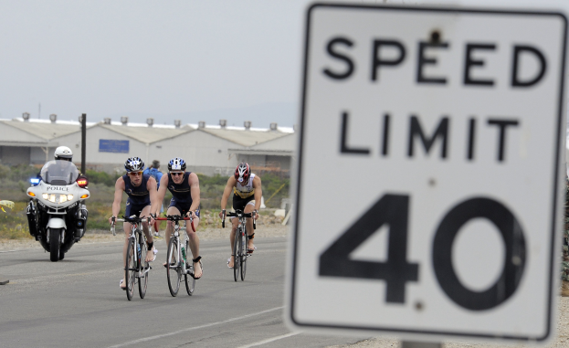 Gruppe von Radfahrern in Helmen, die auf einer Straße neben einem Geschwindigkeitsbegrenzungsschild fahren, mit Gebäuden, Versorgungsmästen, Vegetation und einem klaren Himmel im Hintergrund.