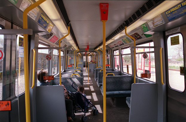 Das Innere eines Zugwagens mit Menschen auf den Sitzreihen, einem Baby im Kinderwagen, offenen Fenstern, die den Blick nach außen freigeben, und Schildern mit Text.