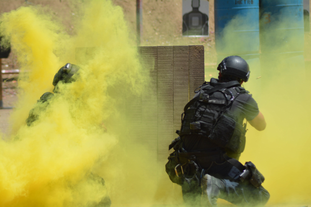 Zwei Polizisten in Einsatzausrüstung mit gelbem Rauch aus dem Mund, mit Helmen und Schutzausrüstung, vor einer Wand mit einer Tafel und anderen Gegenständen, mit dem Boden unten.