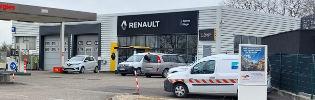 Ein Renault-Autohaus mit parkenden Autos vorne, umgeben von einem Metallzaun, einem Schild, Pfosten, einer Baumgruppe und einem bewölkten Himmel.