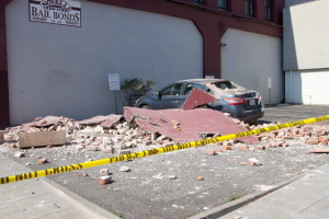 Eine zerbrochene Ziegelwand liegt auf dem Boden mit verstreuten roten Ziegeln, einem geparkten Auto, einer Topfpflanze und einem Gebäude mit einer "Kautionsfreiheit"-Plakette im Hintergrund.
