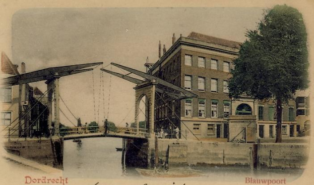 Altes Schwarz-Weiß-Foto einer Brücke unter Bau, die einen Fluss überspannt, mit Gebäuden und Bäumen im Hintergrund und verstreuten Baumaschinen und Materialien an ihrer Basis.