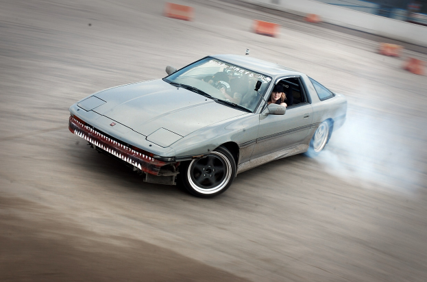 Eine Person in einem Helm fährt ein Auto auf einer Strecke, emitiert Rauch, umgeben von orangenen Objekten, mit einem unscharfen Hintergrund, der hohe Geschwindigkeit andeutet.
