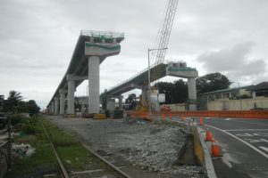 Eine Baustelle mit einer Brücke im Hintergrund, eine Straße mit Absperrbaken auf der rechten Seite, Steine und Gras auf dem Boden, eine Eisenbahnschiene auf der linken Seite, Bäume und Gebäude auf beiden Seiten der Straße und ein bewölkter Himmel.