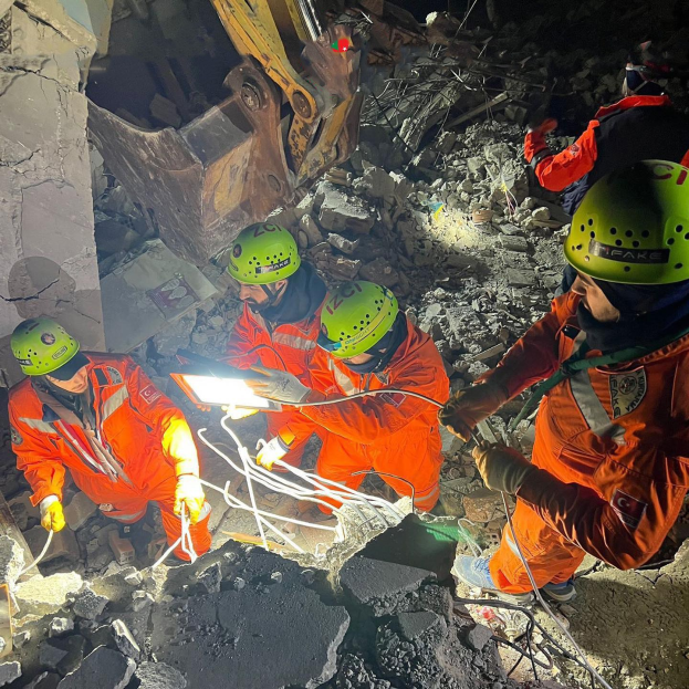 Gruppe von Menschen in orangefarbenen Uniformen und grünen Helmen bei der Arbeit an Schutt mit einem Kran im Hintergrund während einer Rettungsaktion.