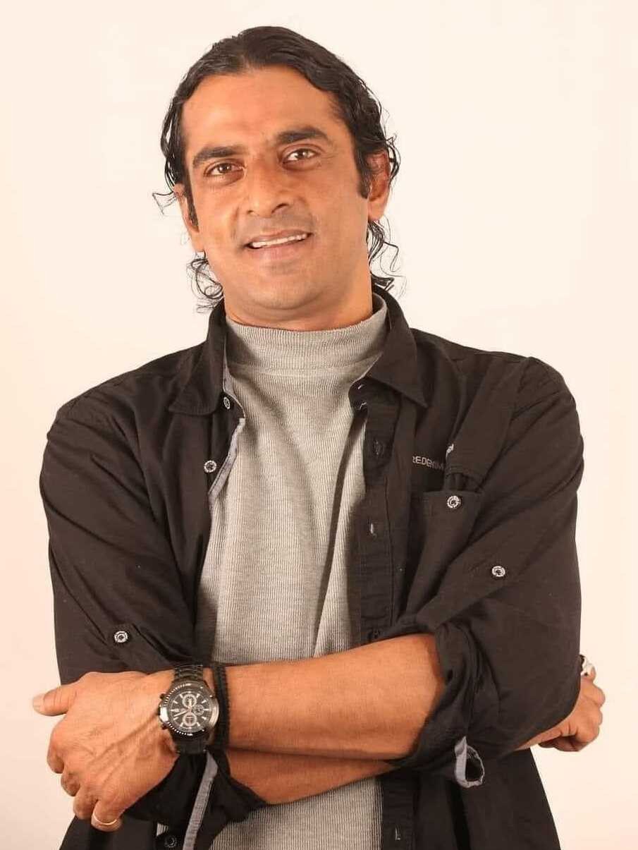 Ein Mann in einem schwarzen und aschfarbenen Anzug mit verschr├Ąnkten Armen, der eine Uhr tr├Ągt und gegen einen weißen Hintergrund steht, der tief nachdenklich wirkt.