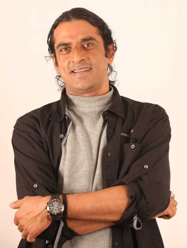 Ein Mann in einem schwarzen und aschfarbenen Anzug mit verschr├Ąnkten Armen, der eine Uhr tr├Ągt und gegen einen weißen Hintergrund steht, der tief nachdenklich wirkt.