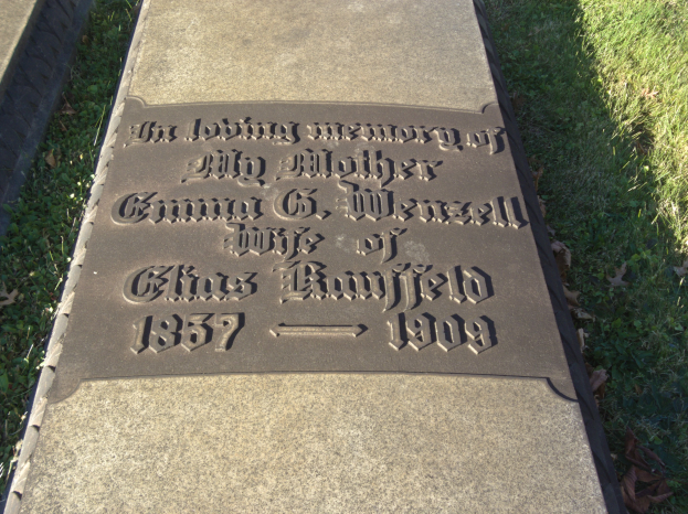 Grabstein auf einem Friedhof mit der Inschrift "Die liebevolle Erinnerung an meine Mutter", umgeben von Gras und welken Blättern.
